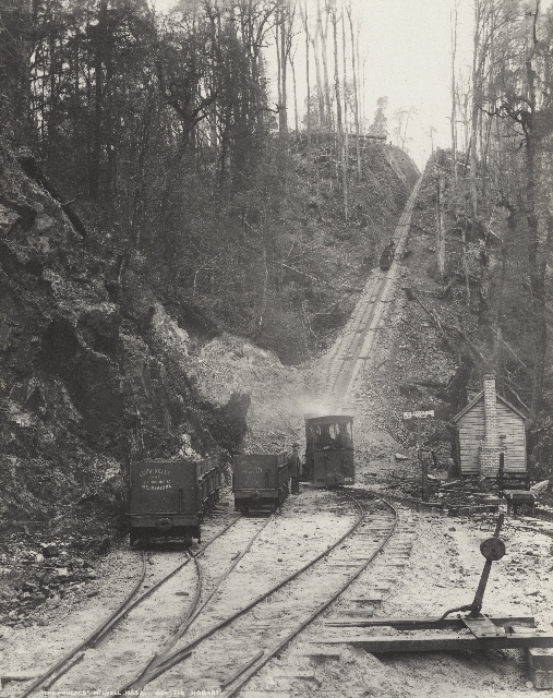 A historic image of a locomotive about to head up a steep section of hill towards Rinadeena. A railway point sits in the foreground and there are several laden carriages behind the loco.