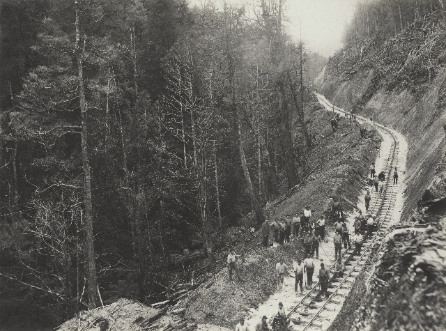 An historical image of track workers working along a section of track with a steep, vertical hill on one side, a flattened section where the track is laid and the hill side dropping off to the left.