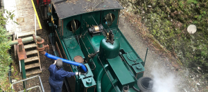 An overhead image of a green steam locomotive being topped up with water through a blue hose. The hose runs from a gravity fed tank at Rinadeena Station.