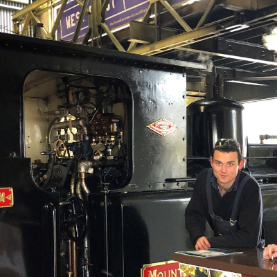 Brock leans on a bar and looks at the camera on the platform at Queenstown Station with Abt Locomotive 3 behind him, 