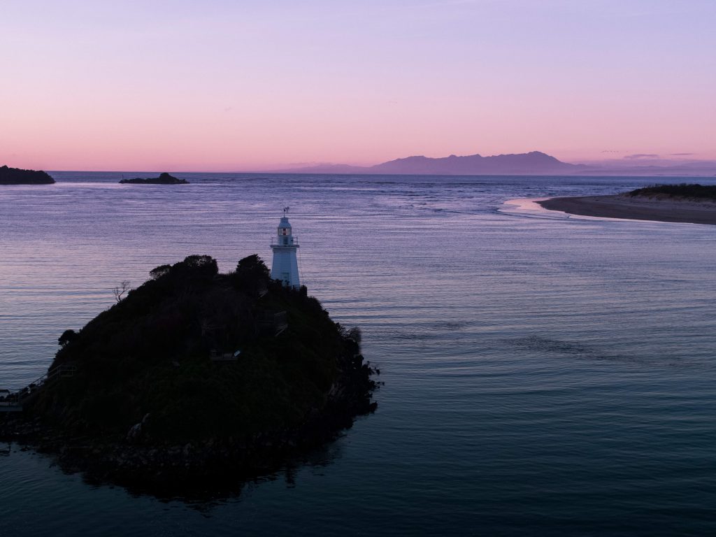 An overhead of the lighthouse at Macquarie Harbour perched on a rocky island. The image is taken at dusk on a calm night and the southern ocean can be seen sitting quietly and calmly in the background.