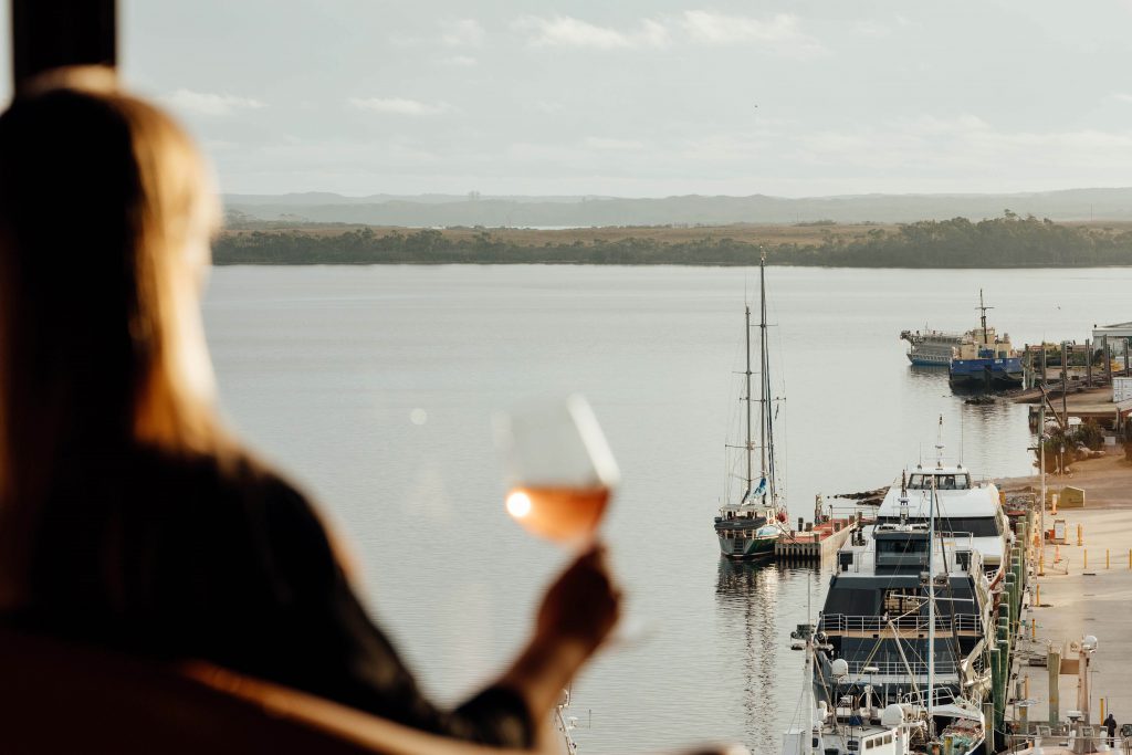 The image is focussed on the waterfront of strahan as taken from a restaurant perched high on the hill. A woman in the foreground is out of focus, but a glass of rose is in her hand and the light shines through it beautifully.