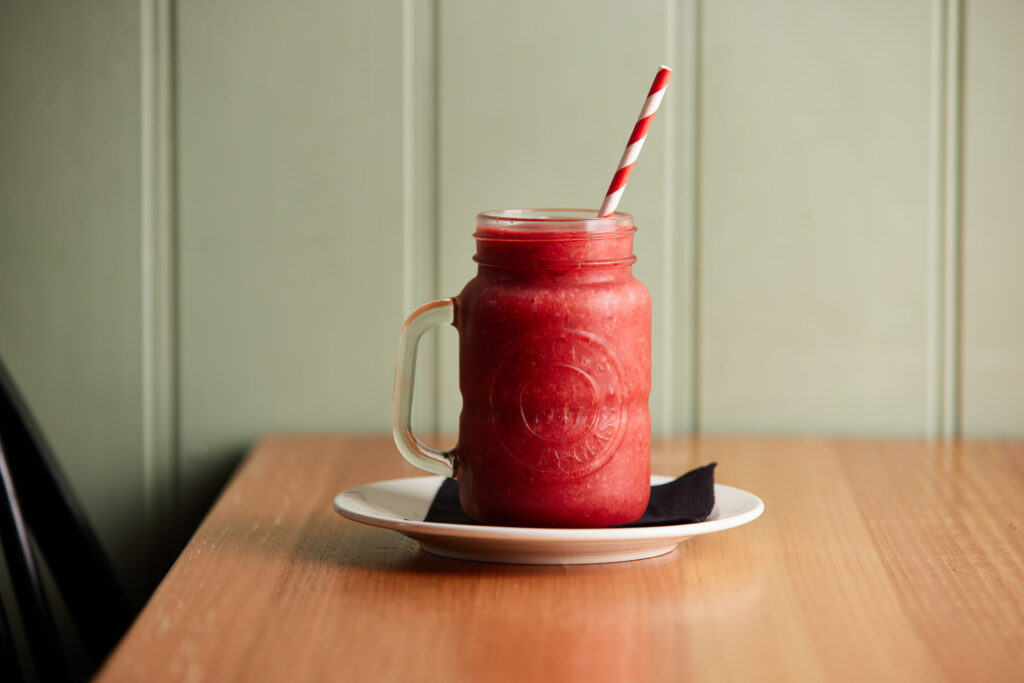 A large glass with a handle sits on a plate with a serviette. Inside the glass is a bright red berry smoothie with red and white paper straw.