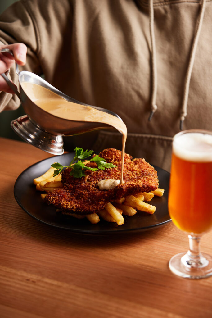 A person in a green jumper pours gravy over a plain chicken schnitzel, sitting on a bed of chips. A glass of beer sits in the foreground.