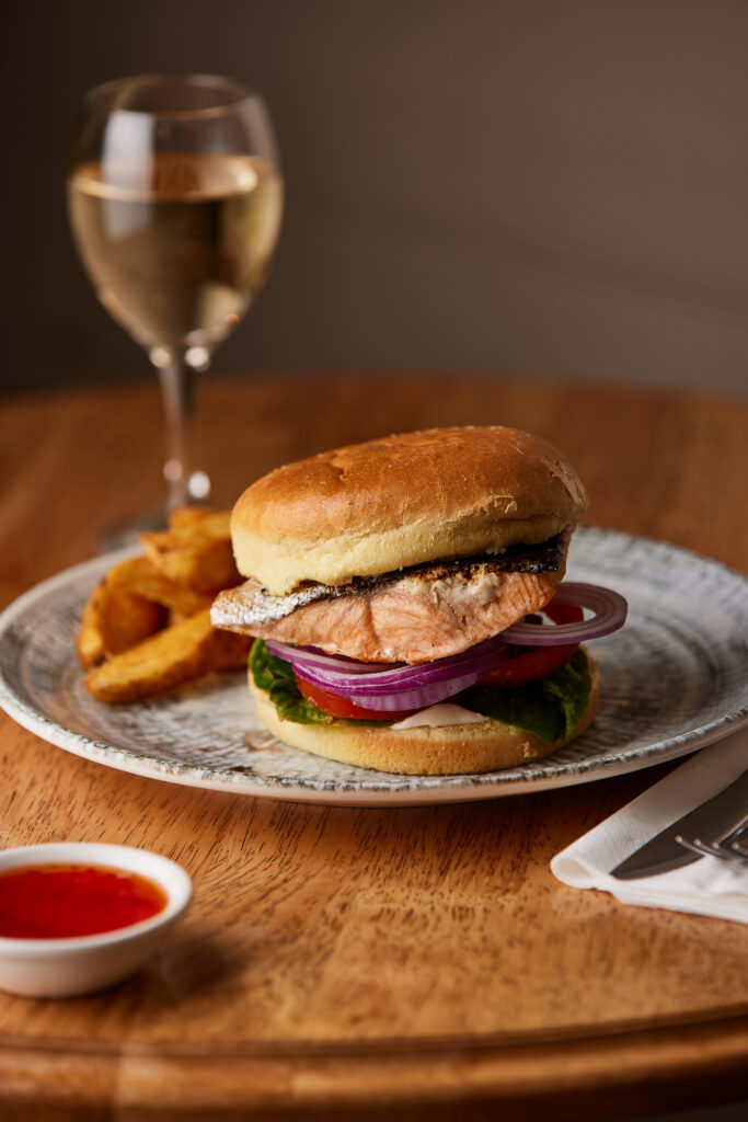 A salmon burger with red onion, lettuce and tomato sits on a plate with some wedges. A glass of wine sits in the background.