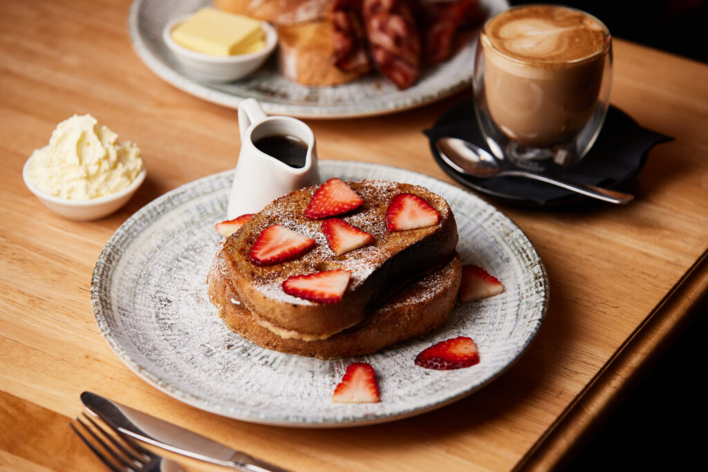 French toast sits on a white and grey decorated plate, topped with strawberries and with a jug of maple syrup on the side.
