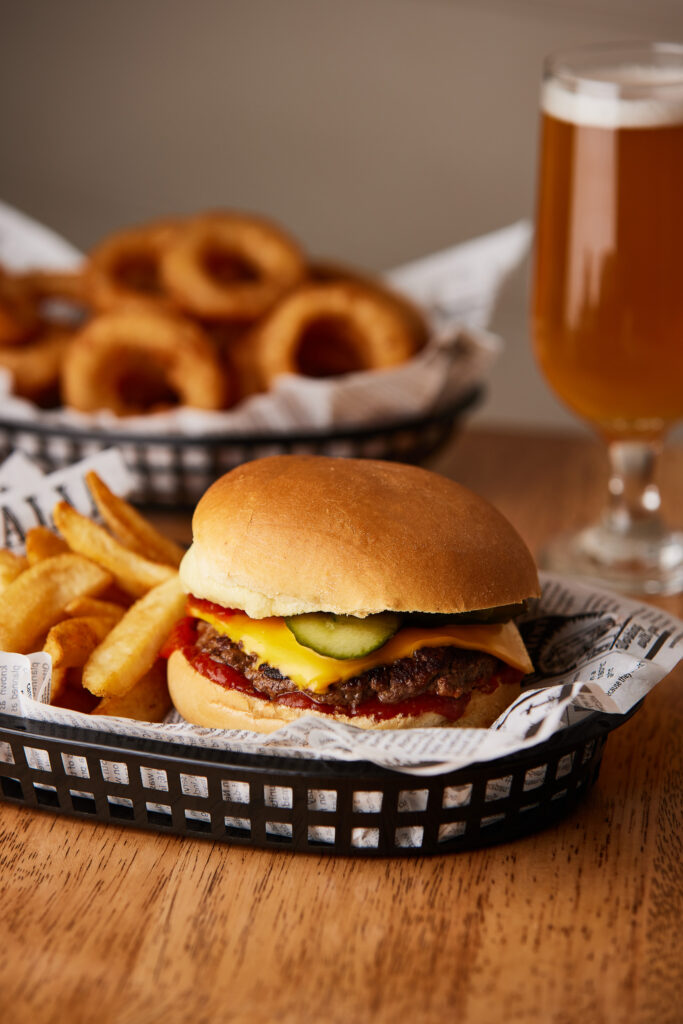 A burger sits in the foreground with onion rings and a glass of beer in the background.