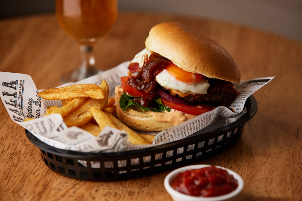 An egg and bacon burger with tomato and lettuce sit in a lined basket with some fries on the side.