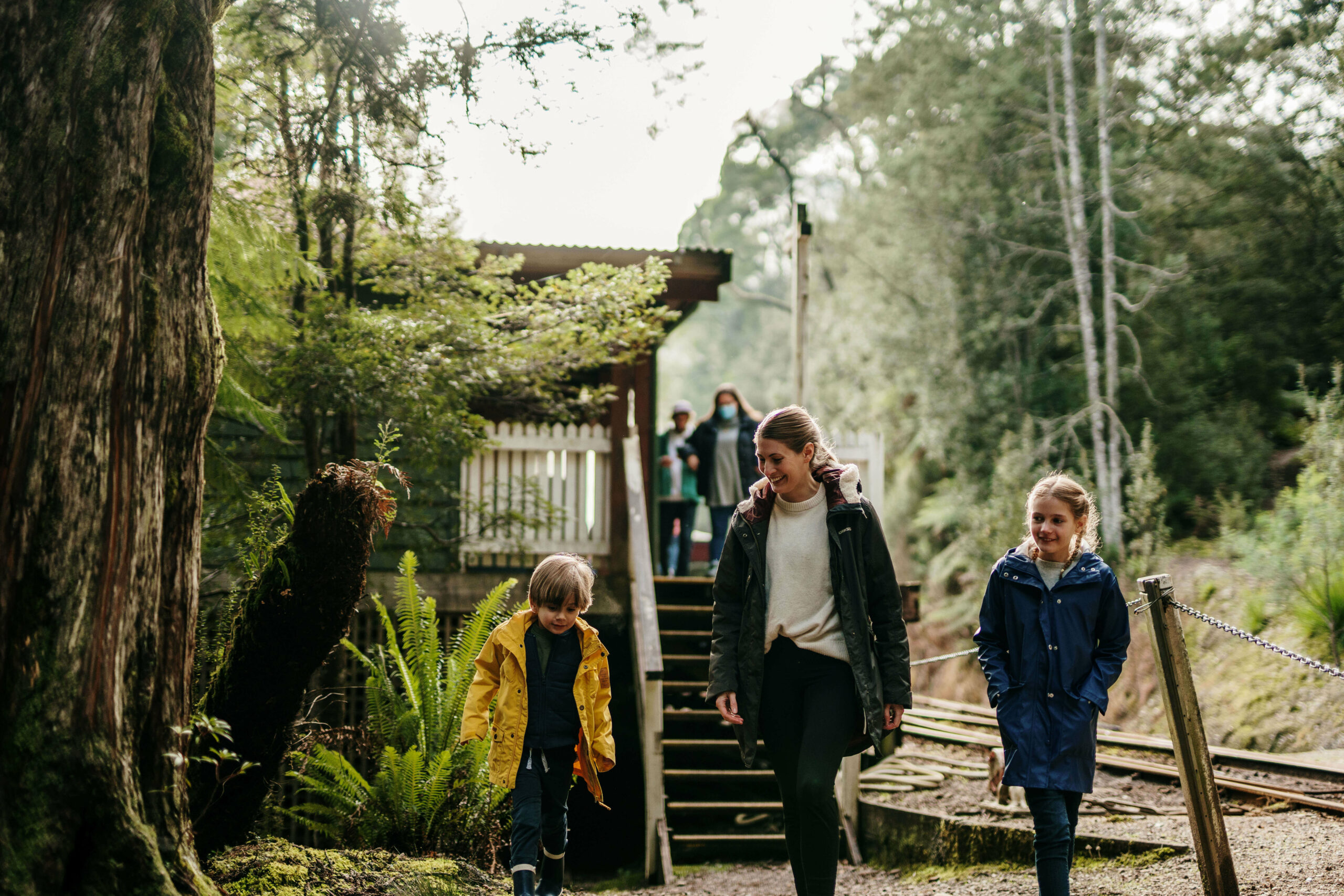A woman wearing a dark jacket and white top accompanies two small children into the rainforest. They are walking away from the platform, which is accessed via wooden stairs. The train track is behind them and to the right.