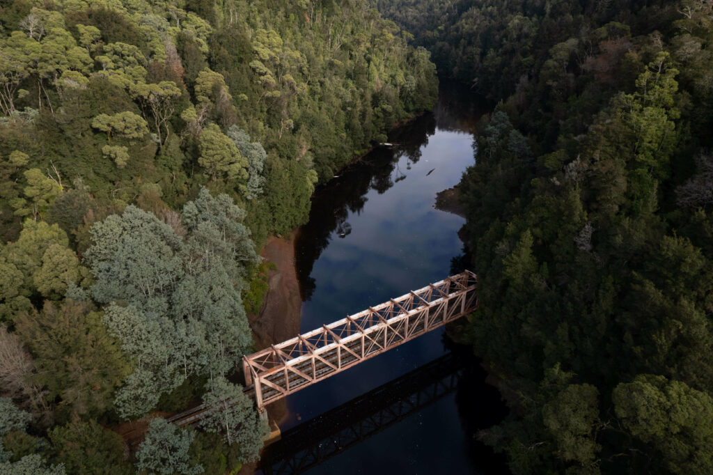 A drone image of the Iron Bridge, which crosses the dark, tannin stained King River. The banks on either side of the river are lush with rainforest growth.
