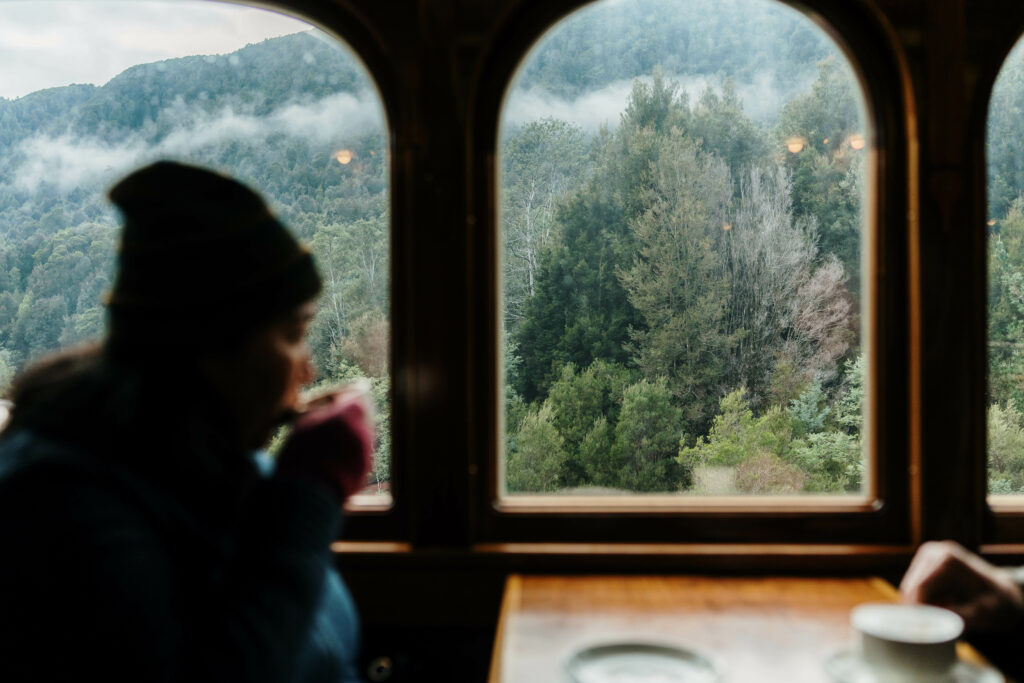 A woman sits on the train sipping a hot drink onboard a wilderness carriage while the rainforest rolls past