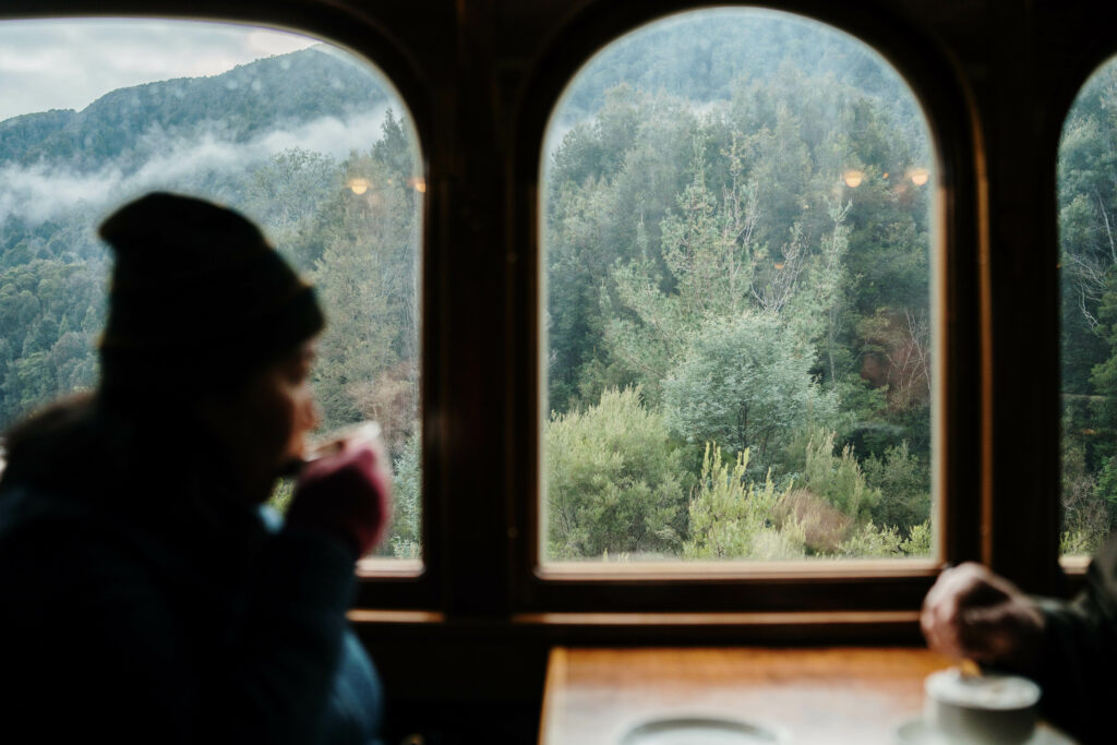 A woman dressed in a woolly jacket and beanie sips at a hot drink onboard a wilderness carriage. A large picture window beside her shows the rainforest in the background.