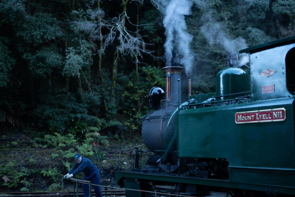 A staff member dressed in blue pushes a green steam locomotive on a manual turntable. The environment is the Tasmanian rainforest in the background.