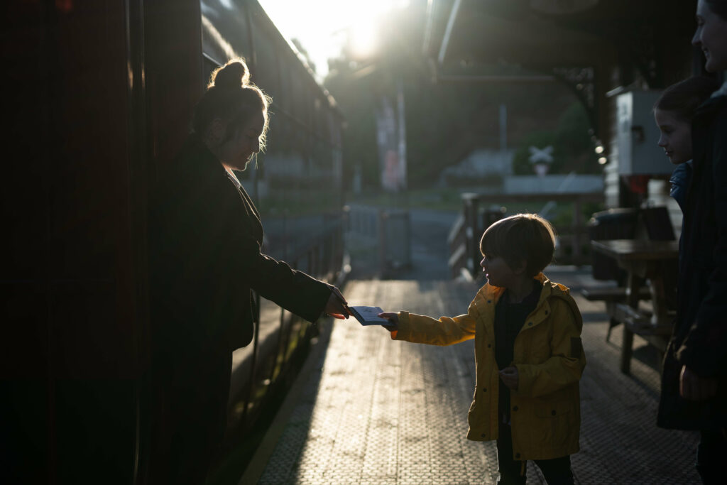 A young boy leans forward on the station platform with an outstretched hand as he hands the steward his ticket for boarding.