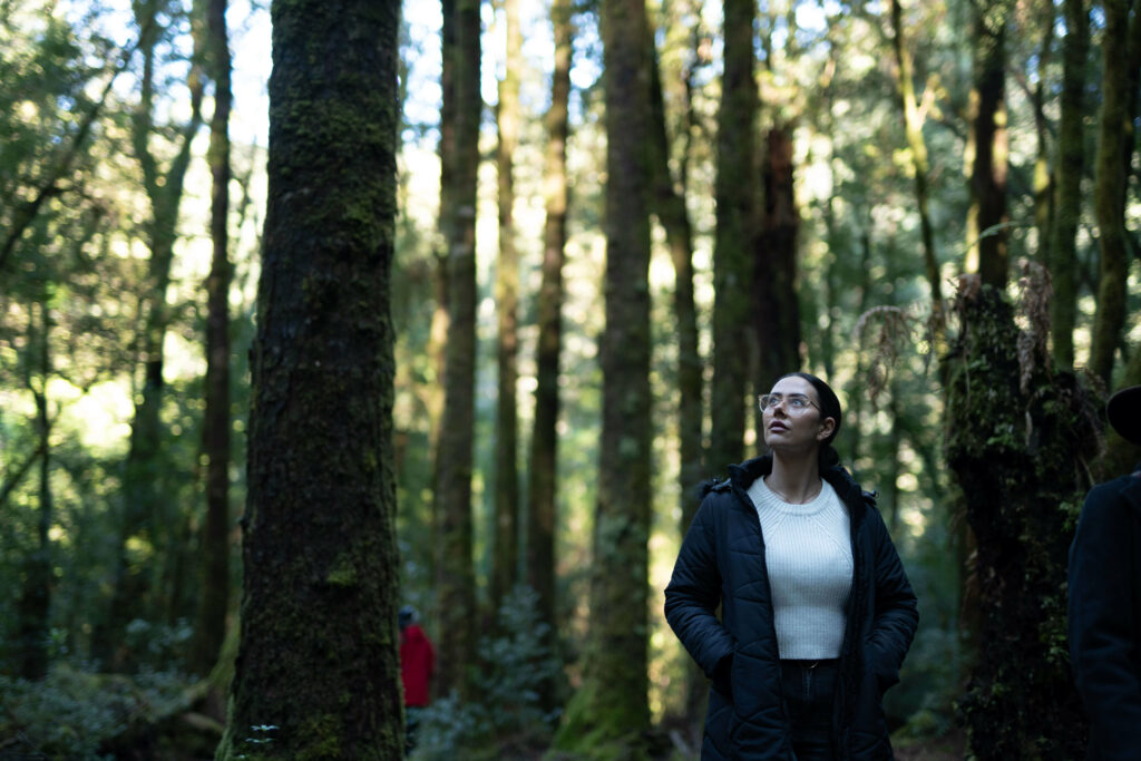 A woman wearing a black parka jacket over a knitted white t-shirt. She wears glasses and is gazing upwards while walking through the rainforest. She is surrounded by tall trees.