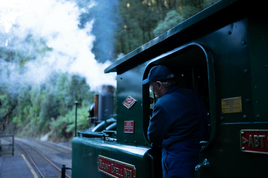 The train driver leans out of the locomotive. He is wearing a blue uniform and cap and peers out at the surrounding rainforest.