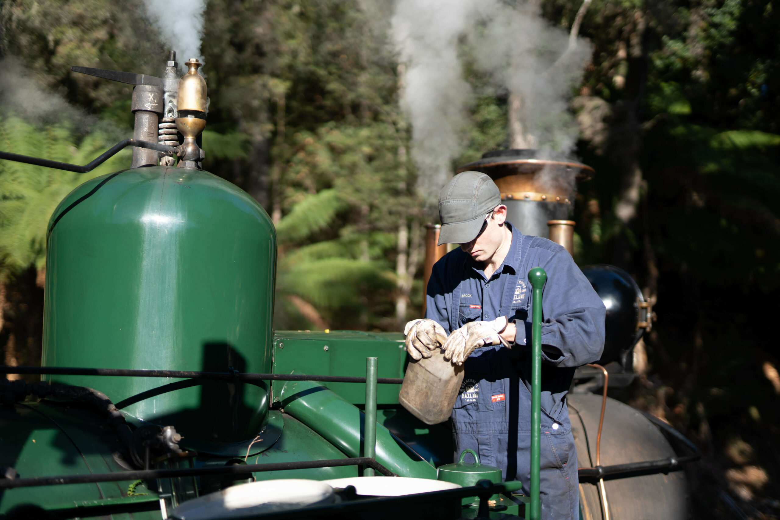 A driver is seen opening a black and greasy oil container while a green locomotive emits steam from brass components to the left of him.