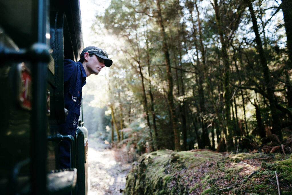 The train driver leans out of the locomotive. He is wearing a blue uniform and cap and peers out at the surrounding rainforest.