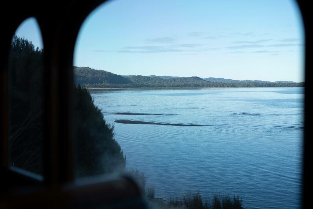 A view of Macquarie Harbour can be seen through the window of the carriage. The water level is low and sand banks can be seen emerging from the water.