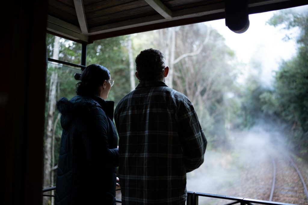 Two passengers enjoy the rainforest behind the train. They are standing on the wilderness carriage balcony.