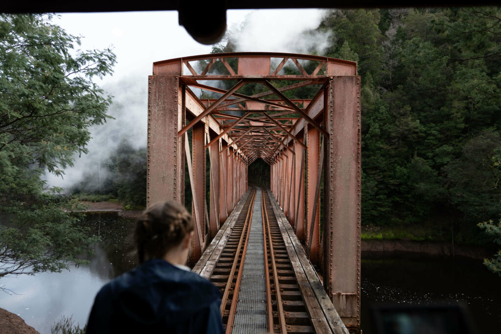 A young girl wearing a blue jacket gazes out behind the train while standing on the carriage balcony. The train has just crossed the King River and can be seen flowing under the Iron Bridge, which is a large reddish steel structure in the background.