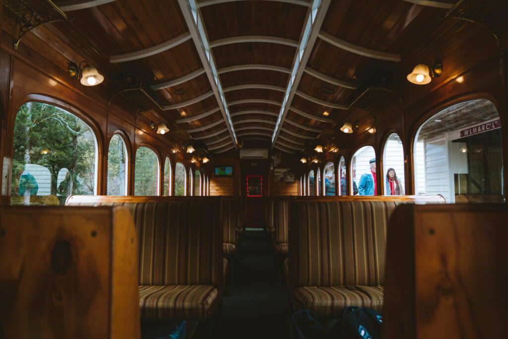 The inside of a darkened heritage carriage sits alongside the platform at Lynchford Station. The carriage contains wooden booth seats with striped upholstery.