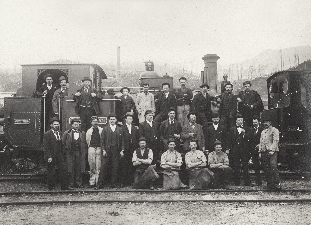 A heritage image of Abt Loco 1 with three lines of Mt Lyell Railway workers in the foreground. The image is black and white and many of the men are wearing suits, while others wear shirts and stained worker trousers.