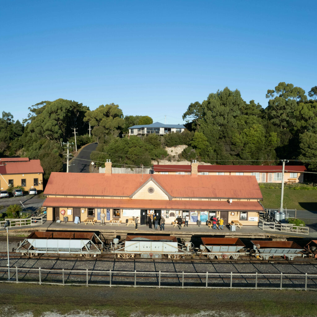 An overhead image of a wooden heritage railway station with a red tin roof. Four open wagons sit on the railway line at the front of the station.