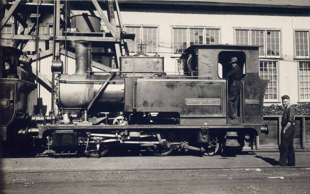 Historical image of Abt loco no. 2 sitting outside original buildings in Queenstown. A man sits inside the cab while another stands in front.