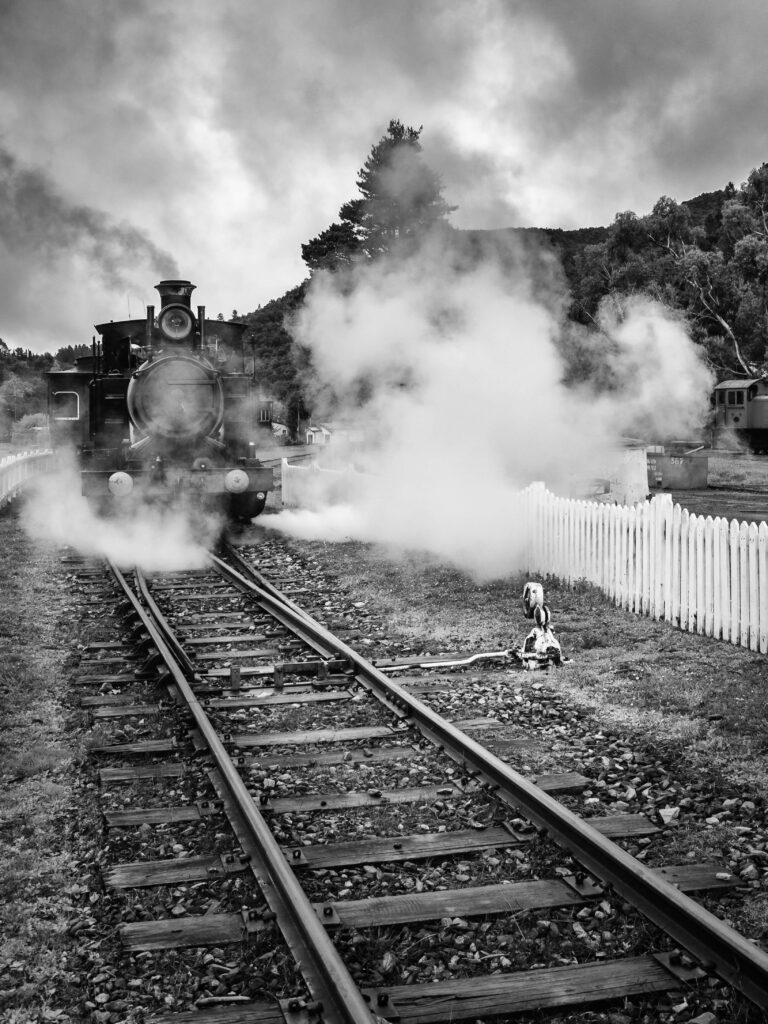 A steam loco emits steam on a railway line outside with a white picket fence running the length of the visible line.