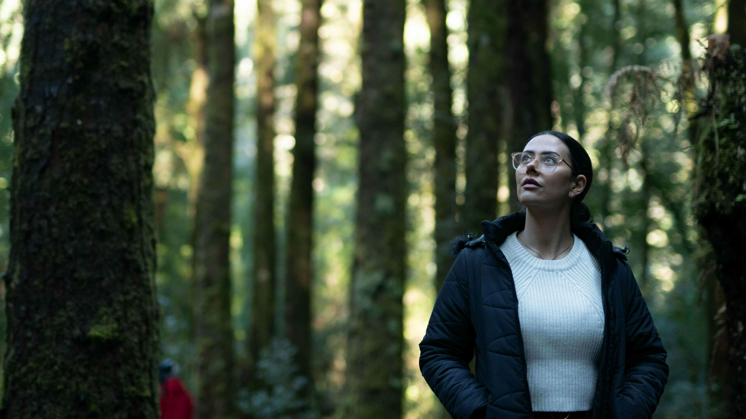 A woman wearing a black parka jacket over a knitted white t-shirt. She wears glasses and is gazing upwards while walking through the rainforest. She is surrounded by tall trees.