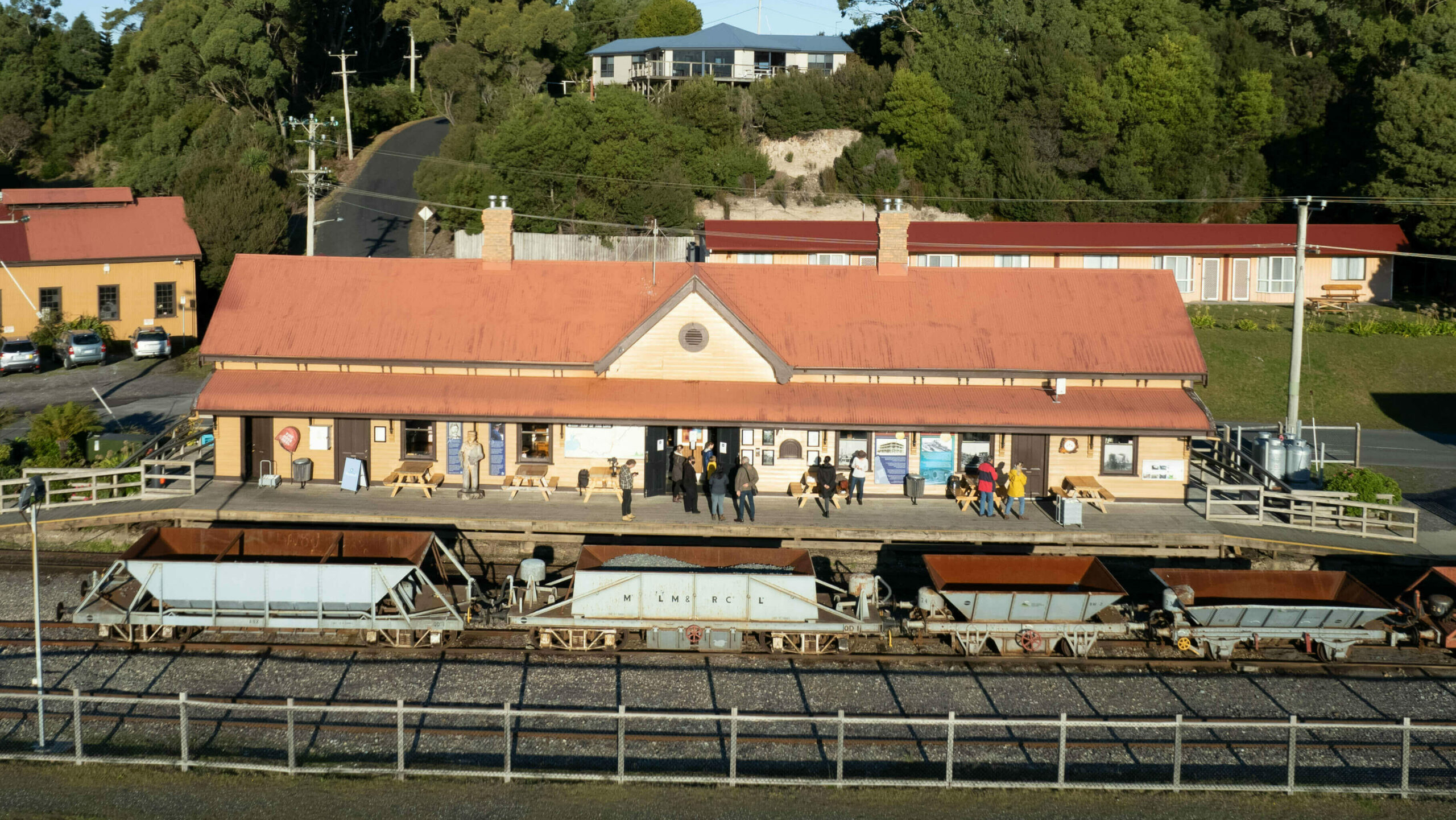 A drone image of Regatta Point Station, now a heritage listed cream coloured wooden building with red tin roof. People mill about on the platform while four open ballust wagons sit alongside the platform.
