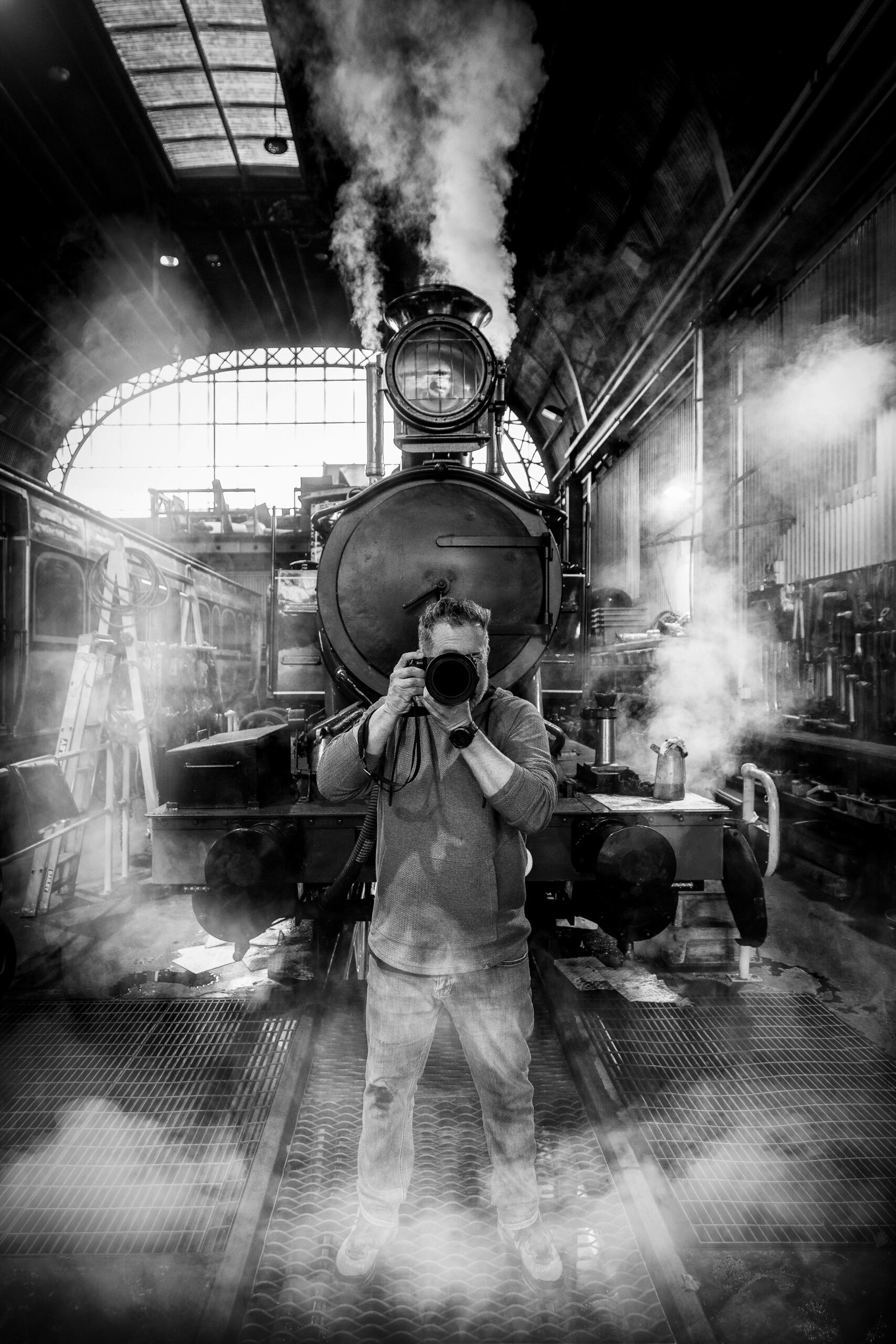 A photographer stands directly in front of a steam locomotive holding a camera in front of his face, facing the direction of the person taking his image.