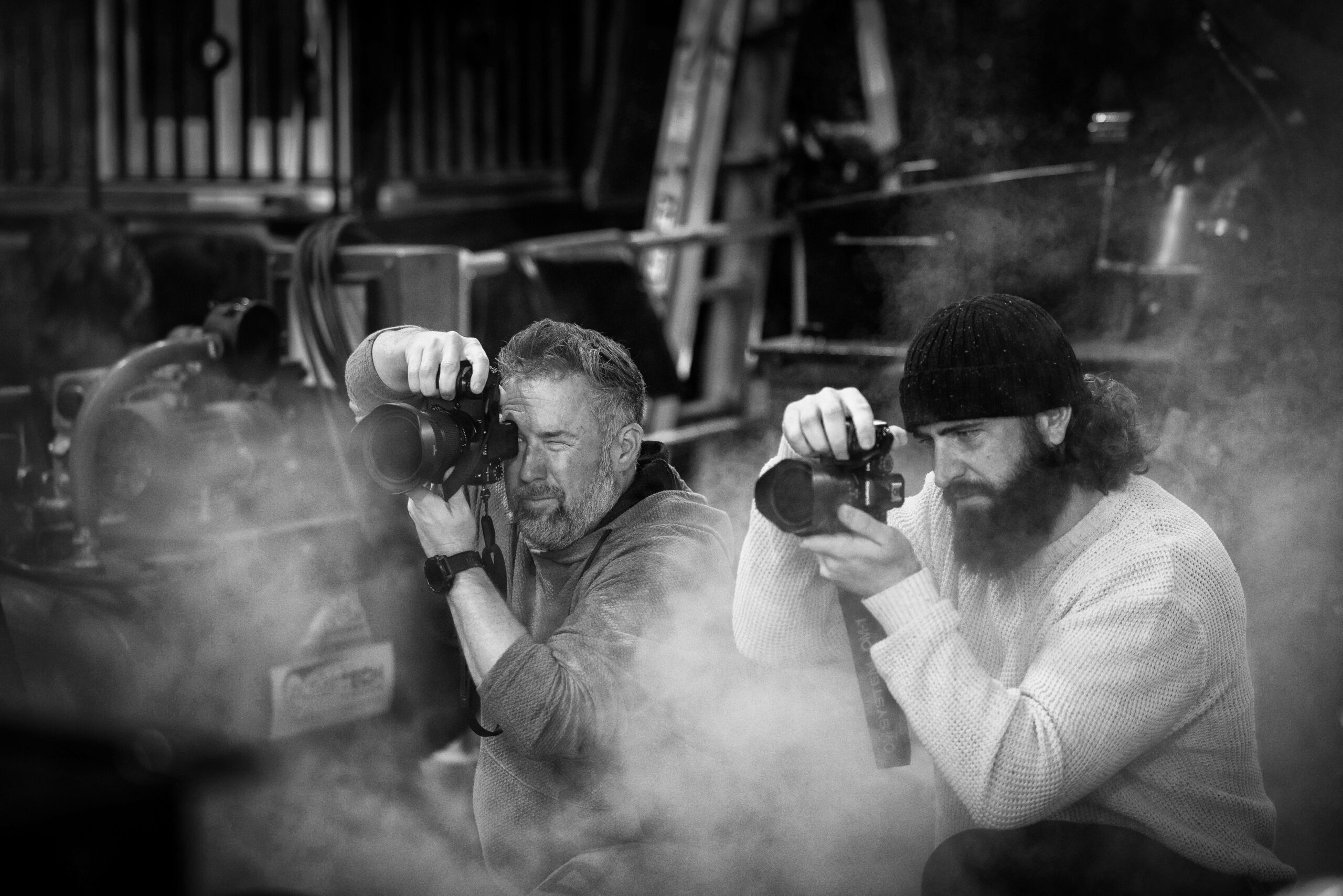 Two men peer into their camera lenses, both surrounded by steam inside the Carswell Park maintenance facility.